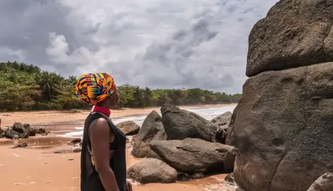 African woman with a colourful headdress and neckpiece standing on a tropical beach looking over the water. 